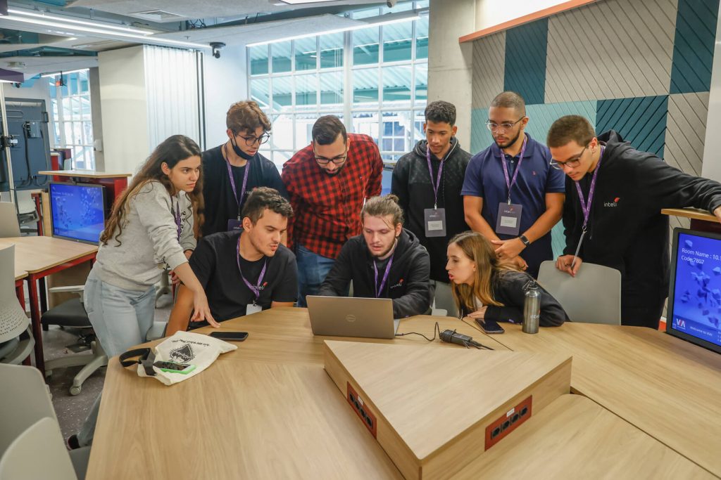 Um grupo de alunos da faculdade Inteli, sentados em mesas de trabalho, colabora em um projeto para uma grande empresa. Eles estão engajados em uma atividade prática, possivelmente brainstorming ou prototipação. A sala é moderna e bem iluminada, com equipamentos e materiais de trabalho visíveis nas mesas. A atmosfera é de concentração e trabalho em equipe, com os alunos interagindo ativamente entre si.