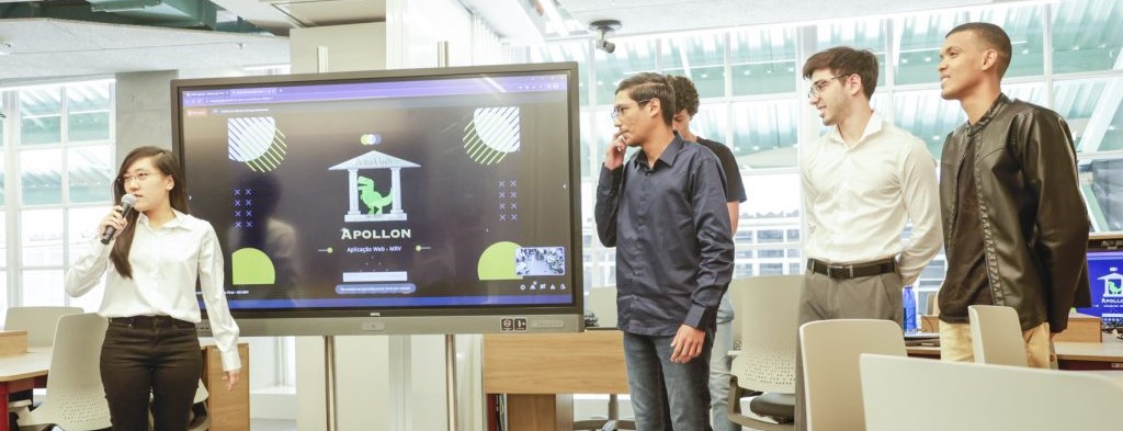 Uma foto horizontal mostra um grupo de alunos do Inteli em uma sala de aula moderna, trabalhando em um projeto para a MRV. Eles estão sentados em mesas, com laptops e materiais de trabalho à sua frente. A atmosfera é de colaboração e foco, com os alunos interagindo e discutindo ideias. Ao fundo, há quadros brancos com anotações e diagramas, indicando o progresso do projeto.